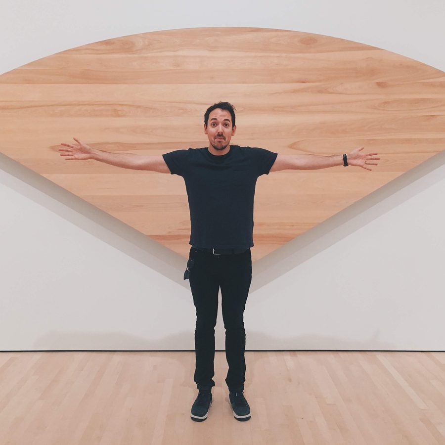 James with arms outstretched in front of a large curved wooden wall sculpture in a museum gallery